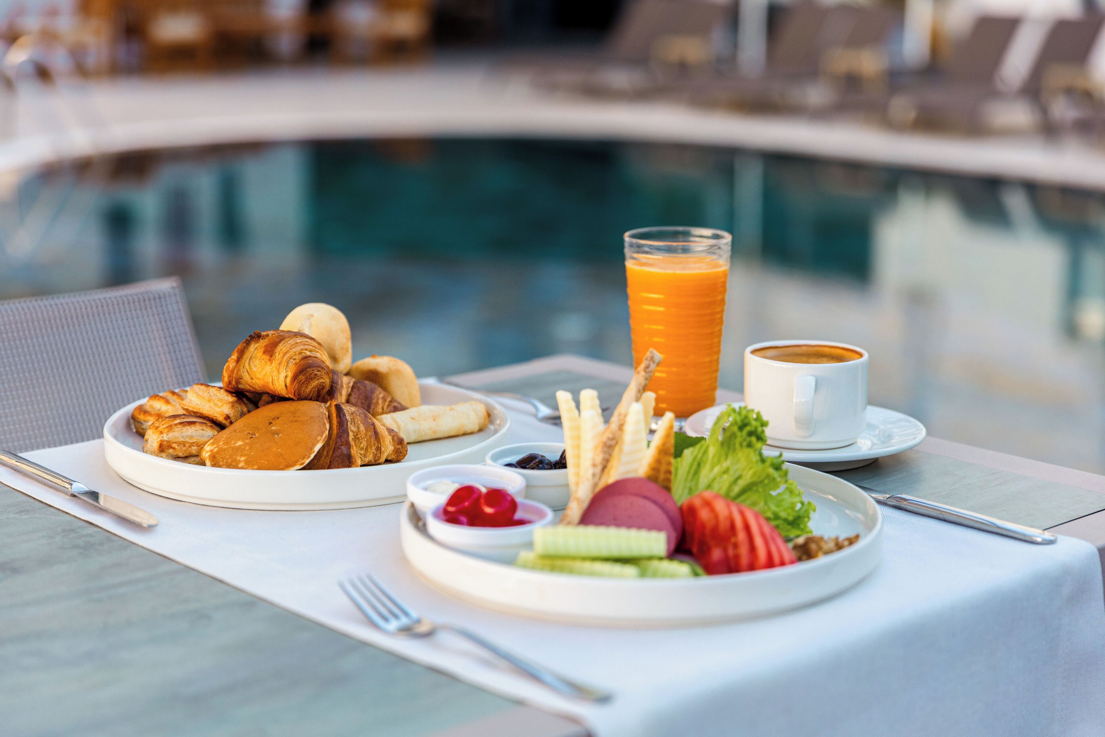 Desayuno con cruasanes, fruta y jugo junto a una piscina.