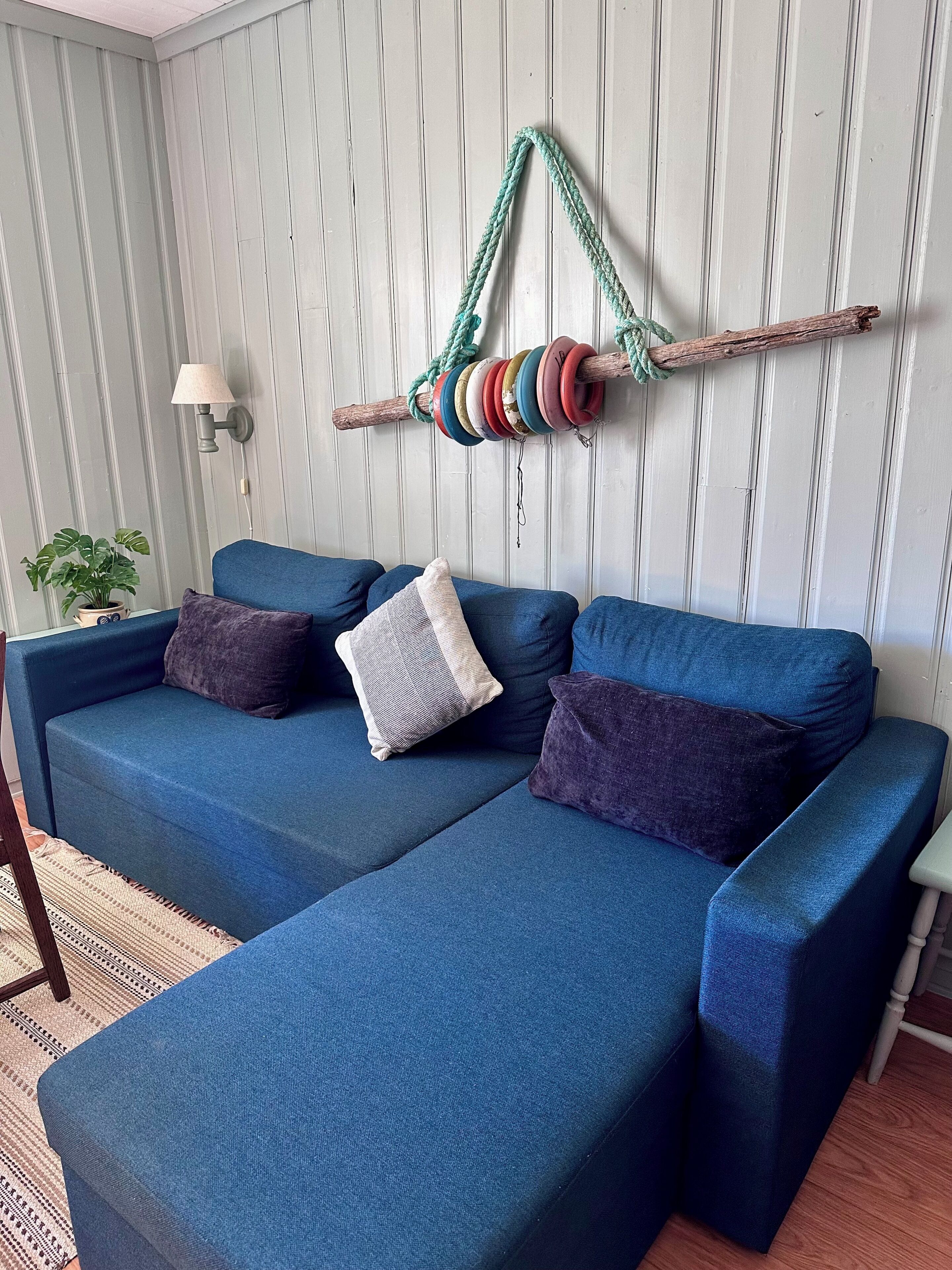 Un sofá seccional azul con cojines, un adorno de cuerda montado en la pared, una planta en maceta y una lámpara.