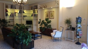 Een woonkamer met een bank, salontafel en potplanten. Er is een boekenkast met boeken en een televisie.
