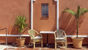 Uma área à beira da piscina, com piso de azulejos, duas cadeiras de vime, duas plantas em vasos e uma janela com moldura branca.