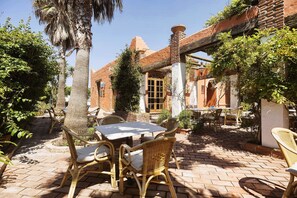An outdoor patio with wicker furniture and a brick floor, surrounded by greenery and a building with a tiled roof. - Hotel Madreselva (Barbate)