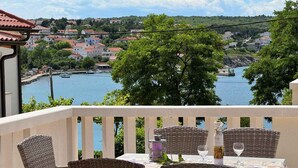 A balcony with wicker furniture and a table set for dining, overlooking a body of water and a town. - Holiday apartment with sea view 20 m from the coast (Šilo)