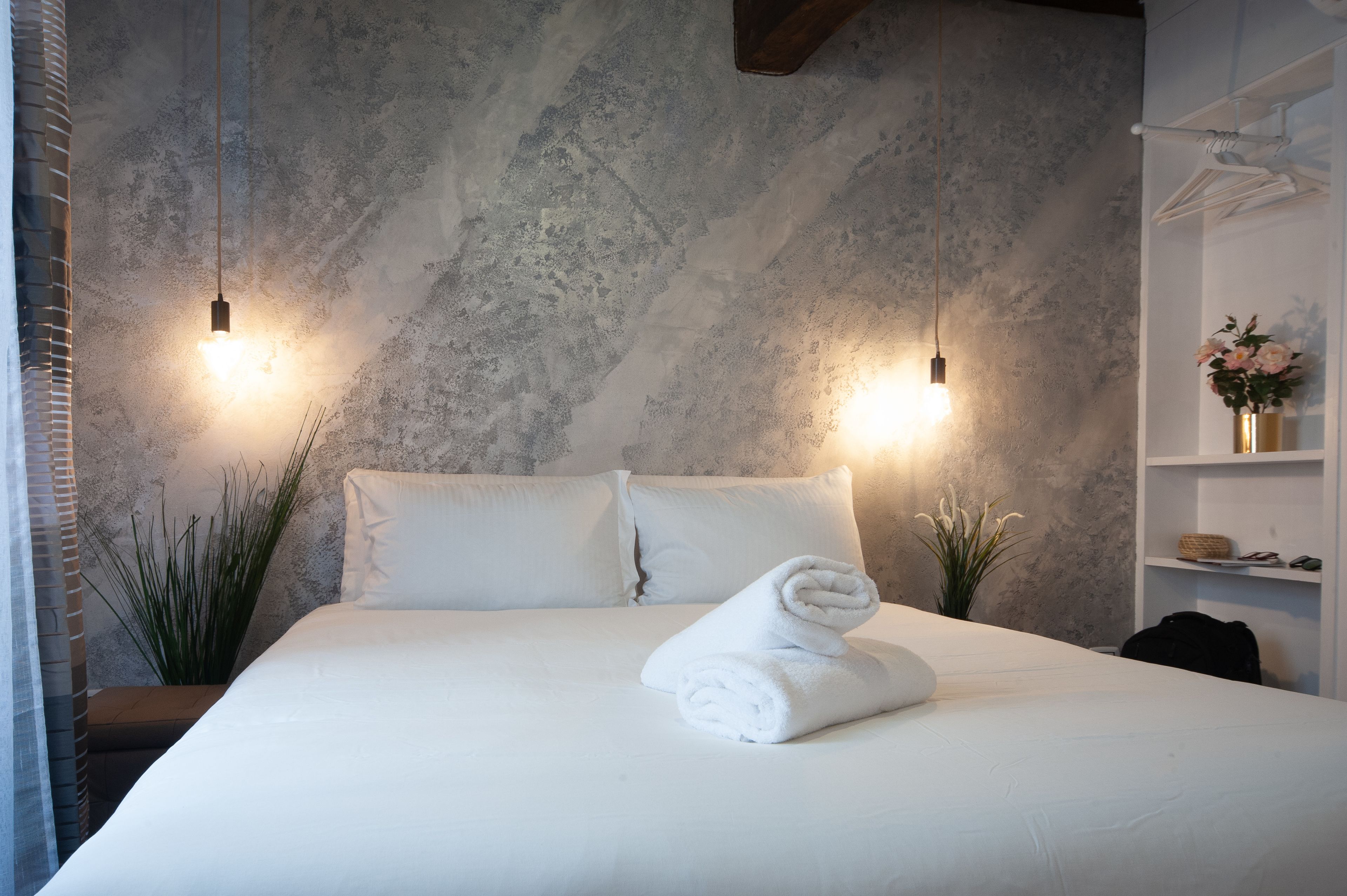 Una cama bien hecha con sábanas blancas y una toalla enrollada, dos luces fijadas en la pared, una planta y un estante con un florero.