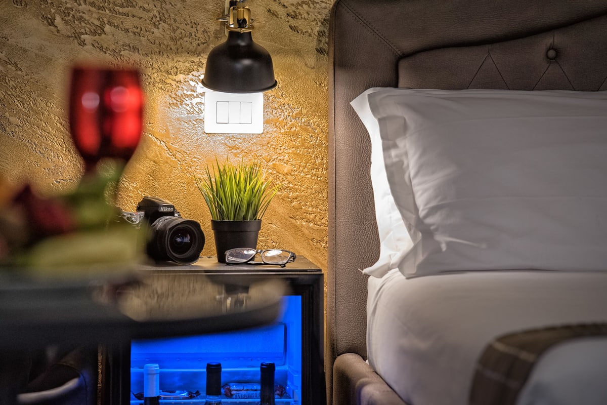 A hotel room with a bed, a nightstand with a lamp, a camera, a plant, and a wall-mounted switch.