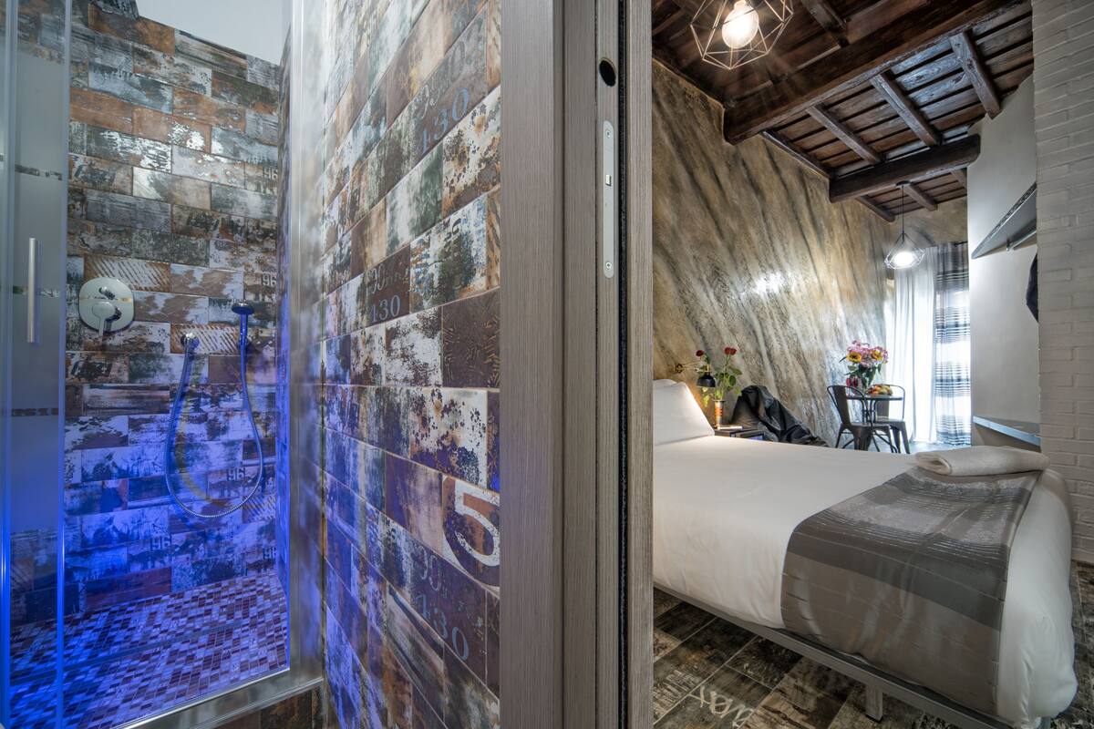 A hotel room with a shower area featuring a unique wall design, a bed with a grey and white striped blanket, and a rustic wooden ceiling.