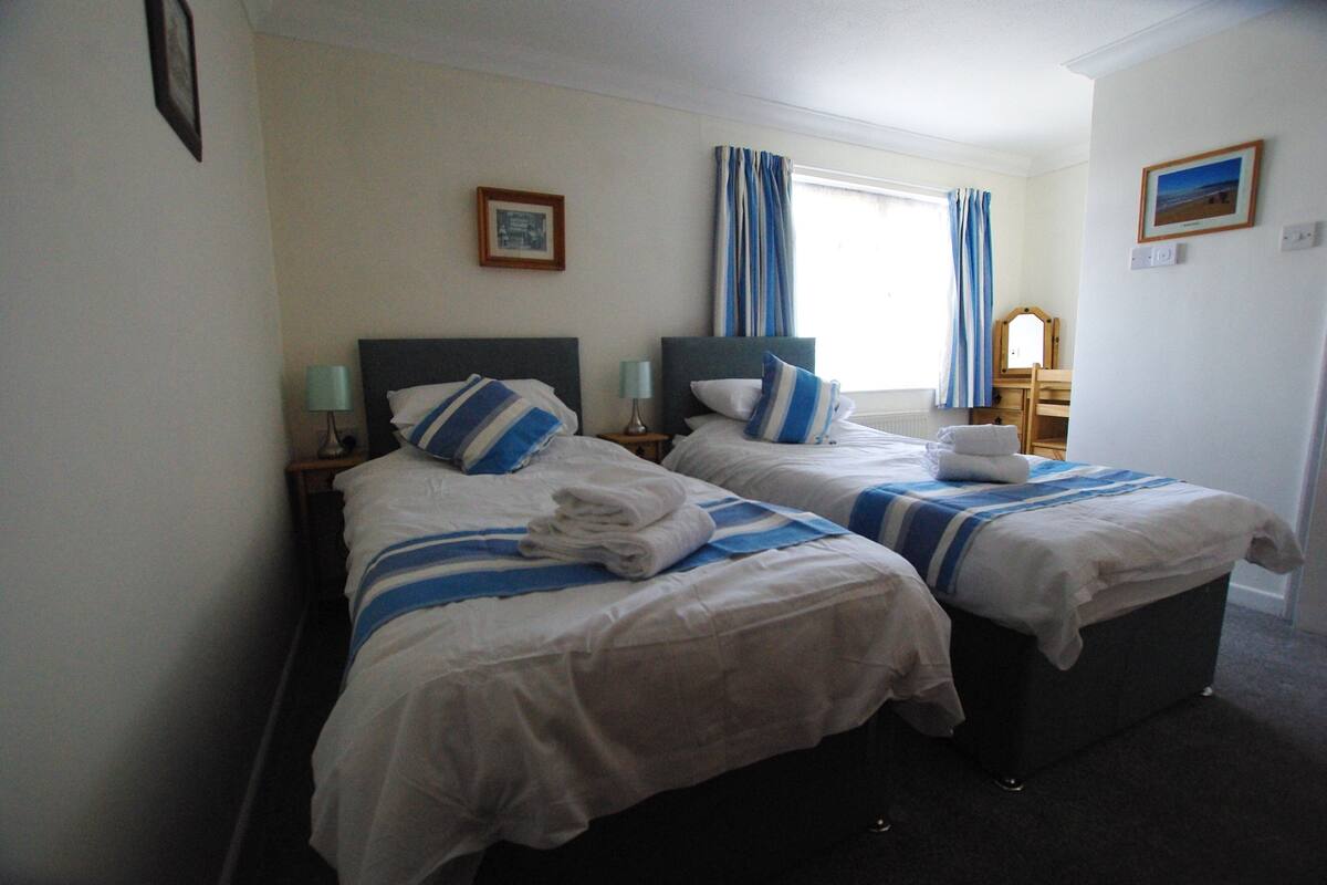 A hotel room with two beds, each with a headboard, a nightstand, a lamp, and a window with curtains.