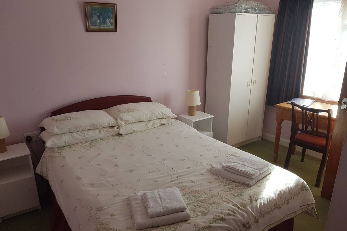 A neatly made bed with floral bedding, a bedside table with a lamp, a wardrobe, and a small desk with a chair.
