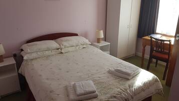 A neatly made bed with floral bedding, a bedside table with a lamp, a wardrobe, and a small desk with a chair.