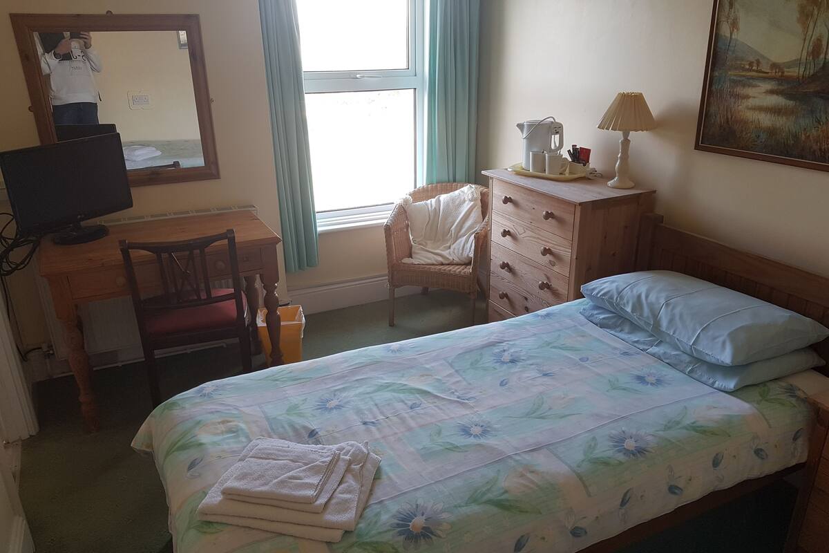 A bedroom with a bed, a desk, a chair, a dresser, a lamp, and a painting on the wall.