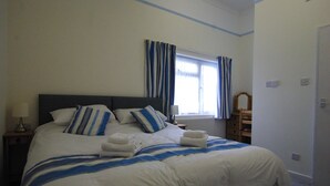 A neatly made bed with white and blue bedding, a bedside table with a lamp, and a window with blue curtains. - Sandhill (Sandown)