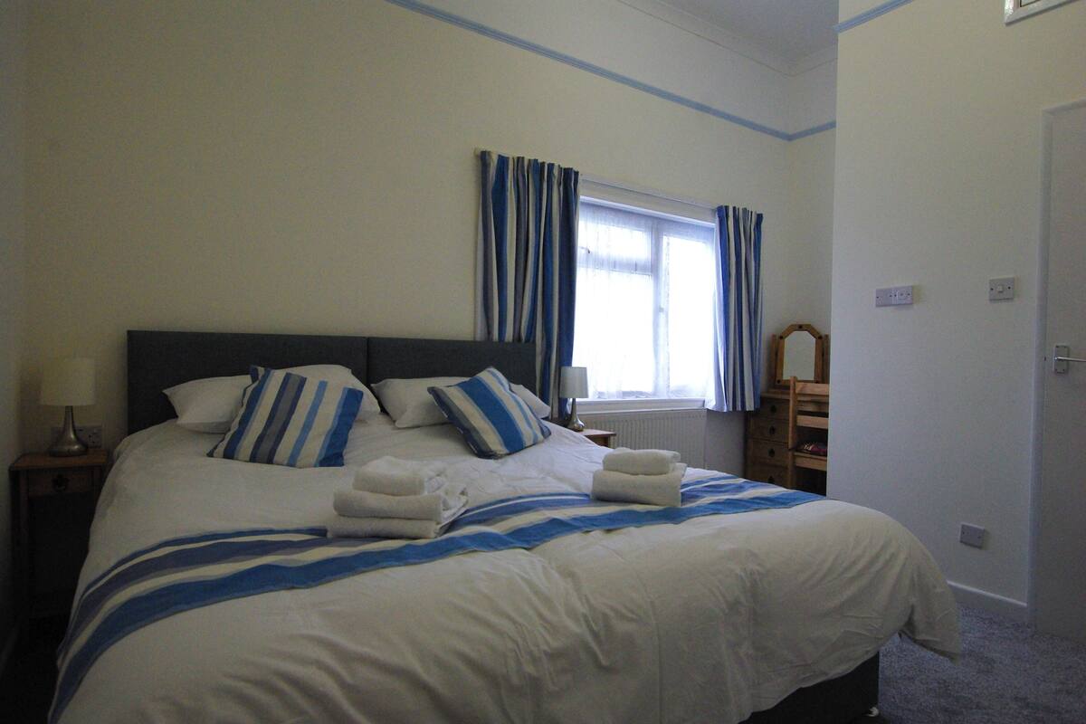 A neatly made bed with white and blue bedding, a bedside table with a lamp, and a window with blue curtains.