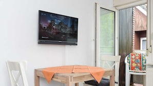 A room with a wooden table and two chairs, a television mounted on the wall, and a sliding glass door leading to an outdoor area.