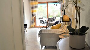 Una sala de estar con un sofá, una mesa de comedor y sillas. Hay una ventana con cortinas, una planta en maceta y una lámpara.