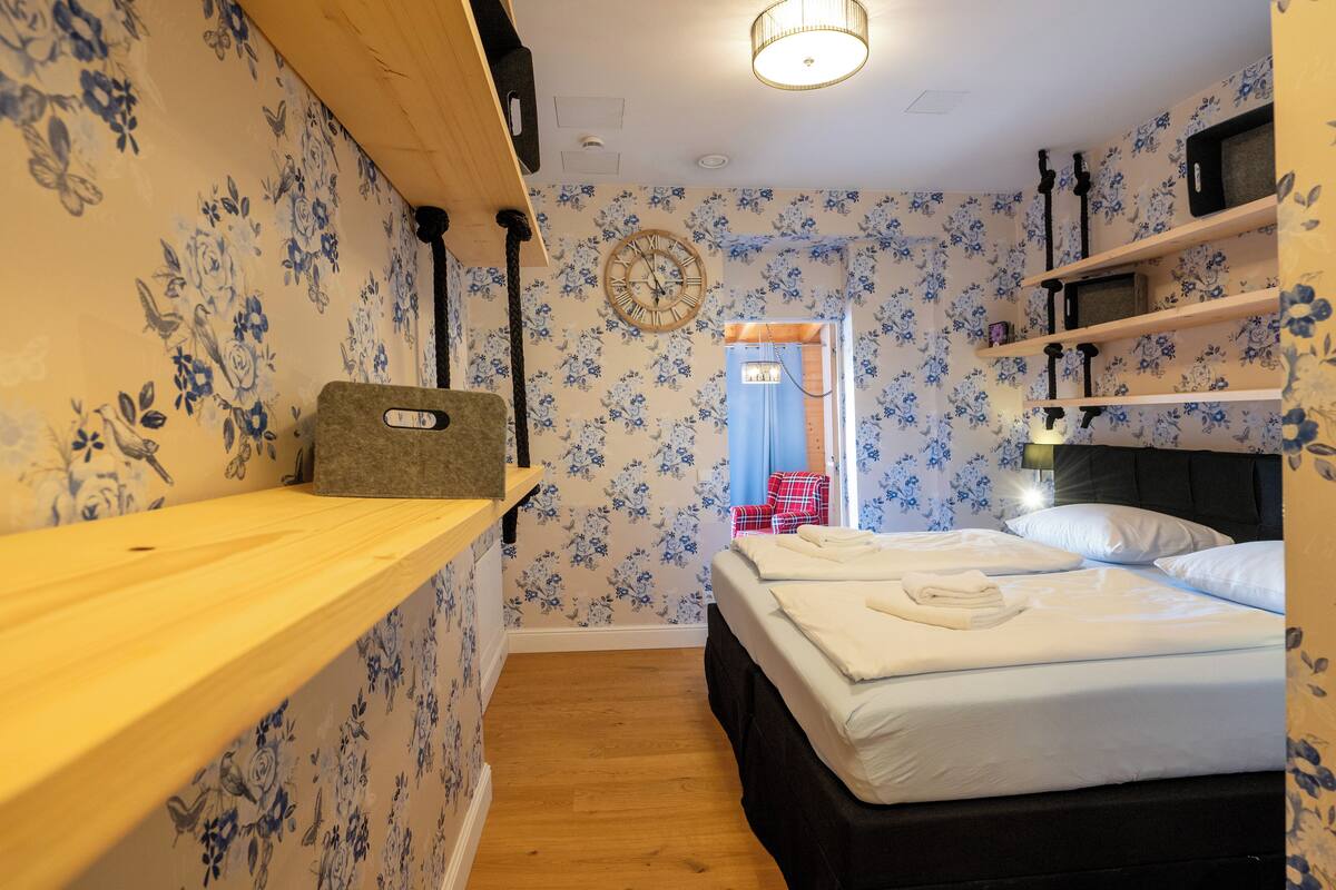 A bedroom with floral wallpaper, a wooden shelf, a bed with white linens, and a clock on the wall.