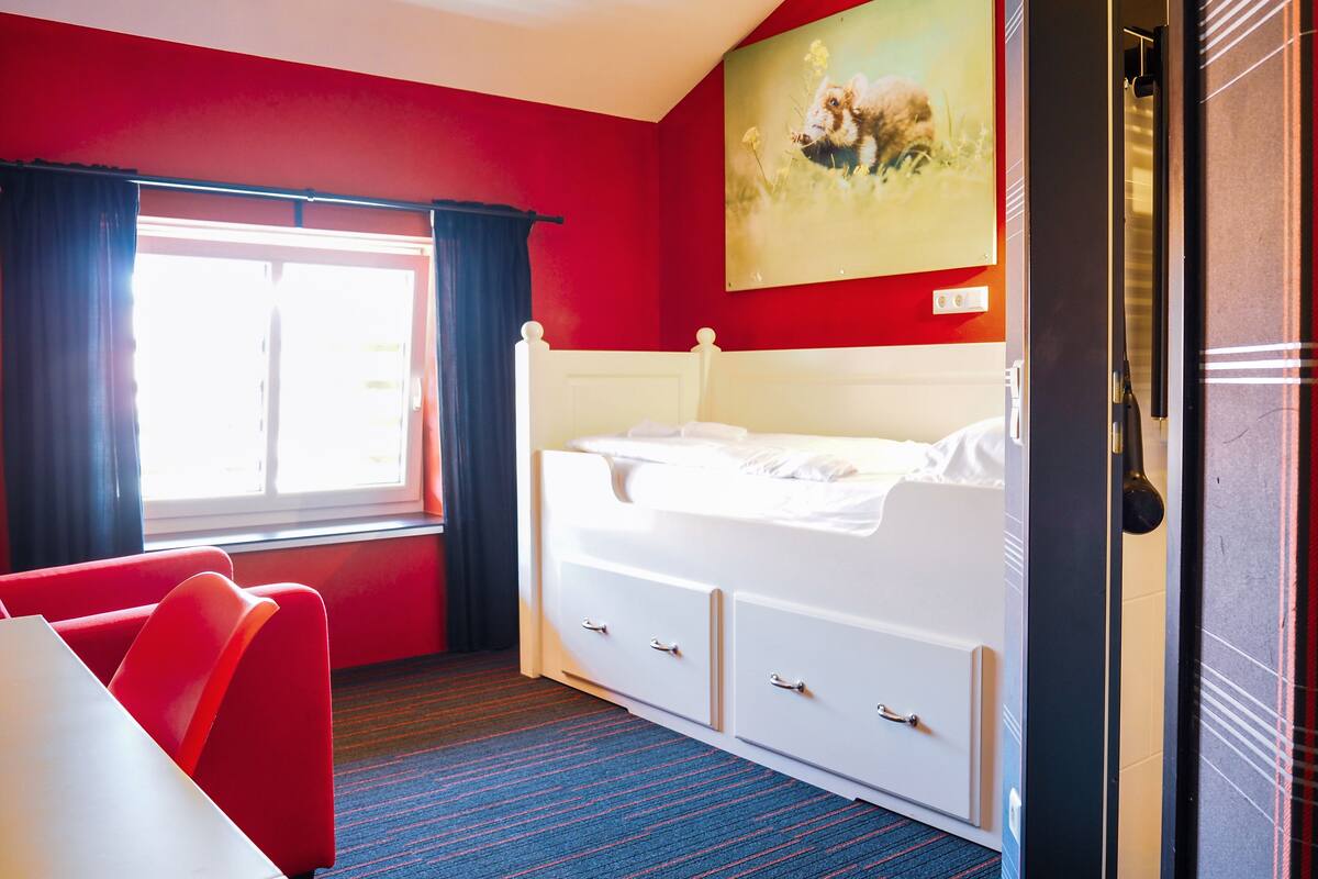 A hotel room with a red wall, a white bed, a red armchair, and a window with curtains.