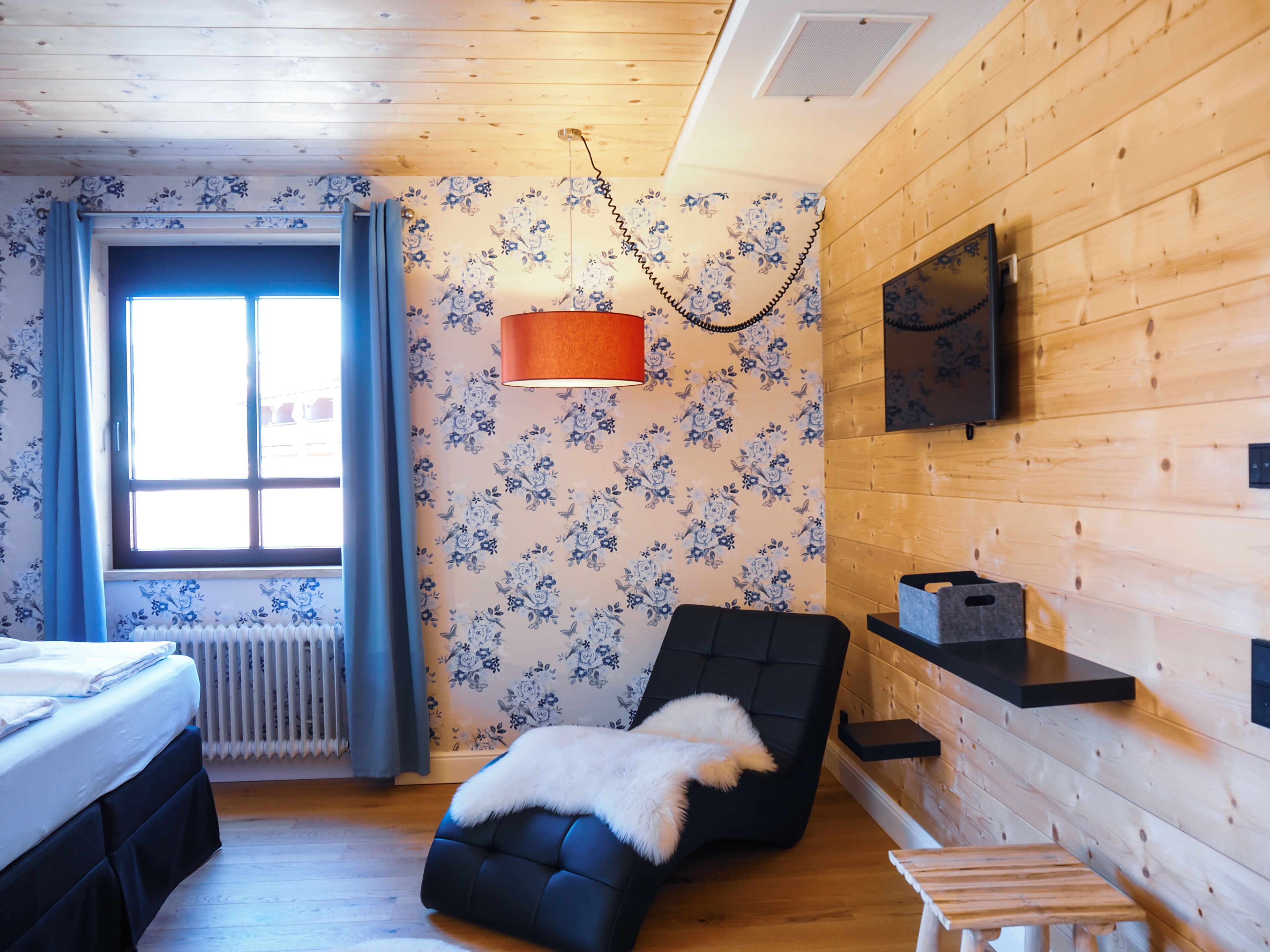 A cozy room with a bed, a black chair with a fur throw, a wooden floor, and a window with curtains.