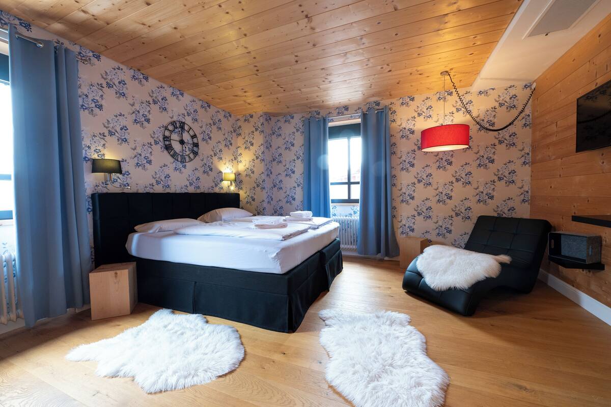 A bedroom with a wooden ceiling, floral wallpaper, a bed with a black headboard, a white fur rug, a black chair with a white fur throw, and a red pendant light.