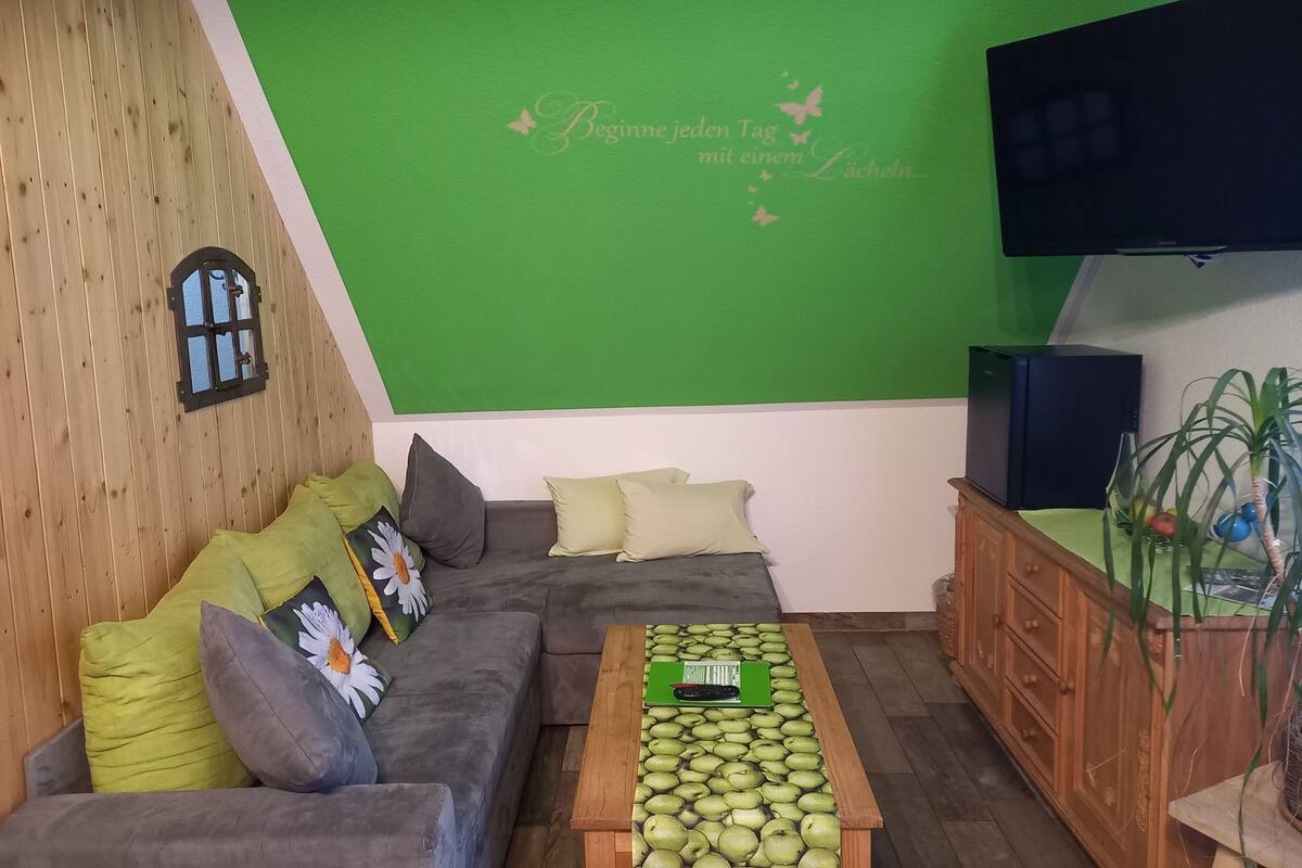 A cozy living room with a green accent wall, a wooden ceiling, a grey sofa with cushions, a wooden coffee table, and a flat-screen TV.