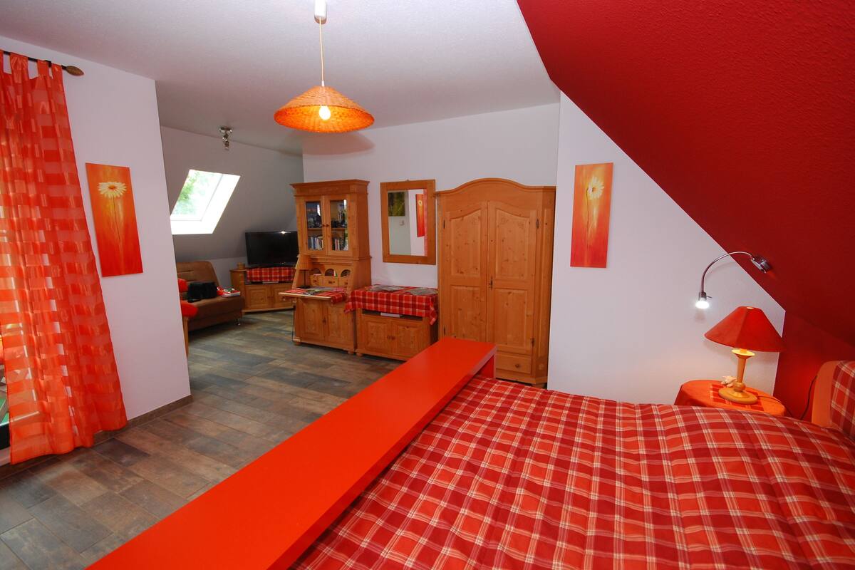 A bedroom with a bed, a wooden wardrobe, a red sofa, and a television.