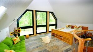 Una habitación con un sofá verde, una cama con almohadillas estampadas, suelo de madera y ventanas amplias con vistas a árboles.
