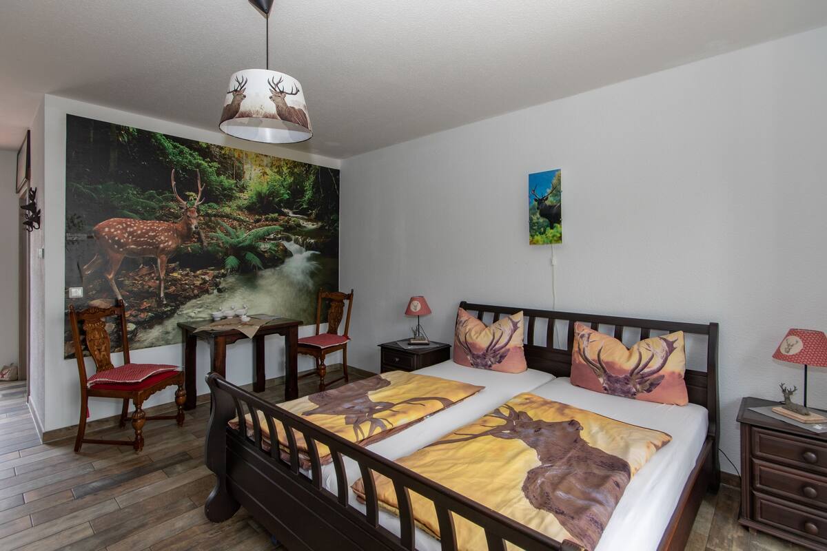 A bedroom with a large forest-themed mural, a double bed with patterned bedding, a wooden nightstand, a chair, and a lamp.