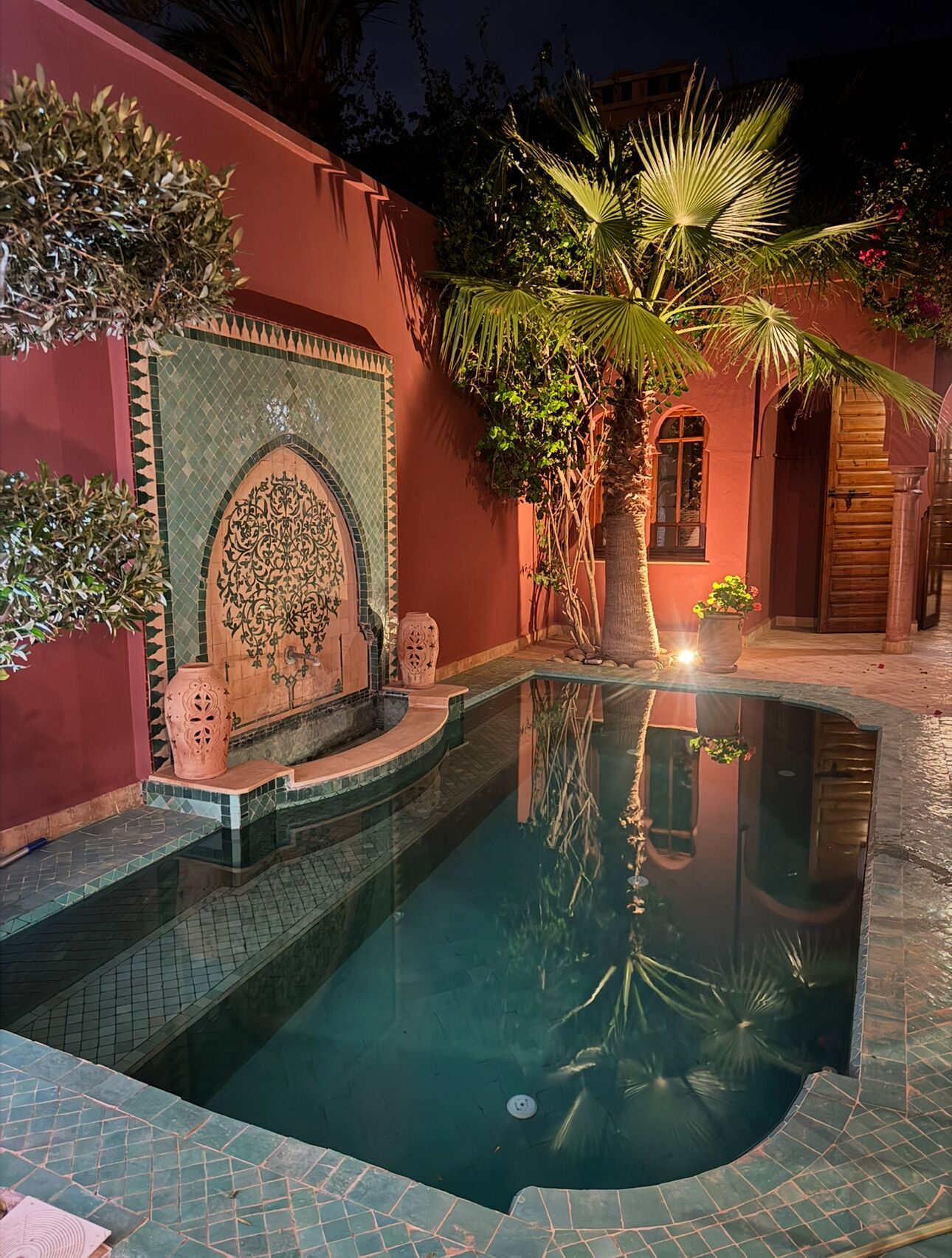 Una piscina con borde de azulejos, rodeada por un muro rojo con un diseño de mosaicos decorativos y una palmera.