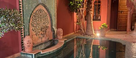 Una piscina con borde de azulejos, rodeada por un muro rojo con un diseño de mosaicos decorativos y una palmera.