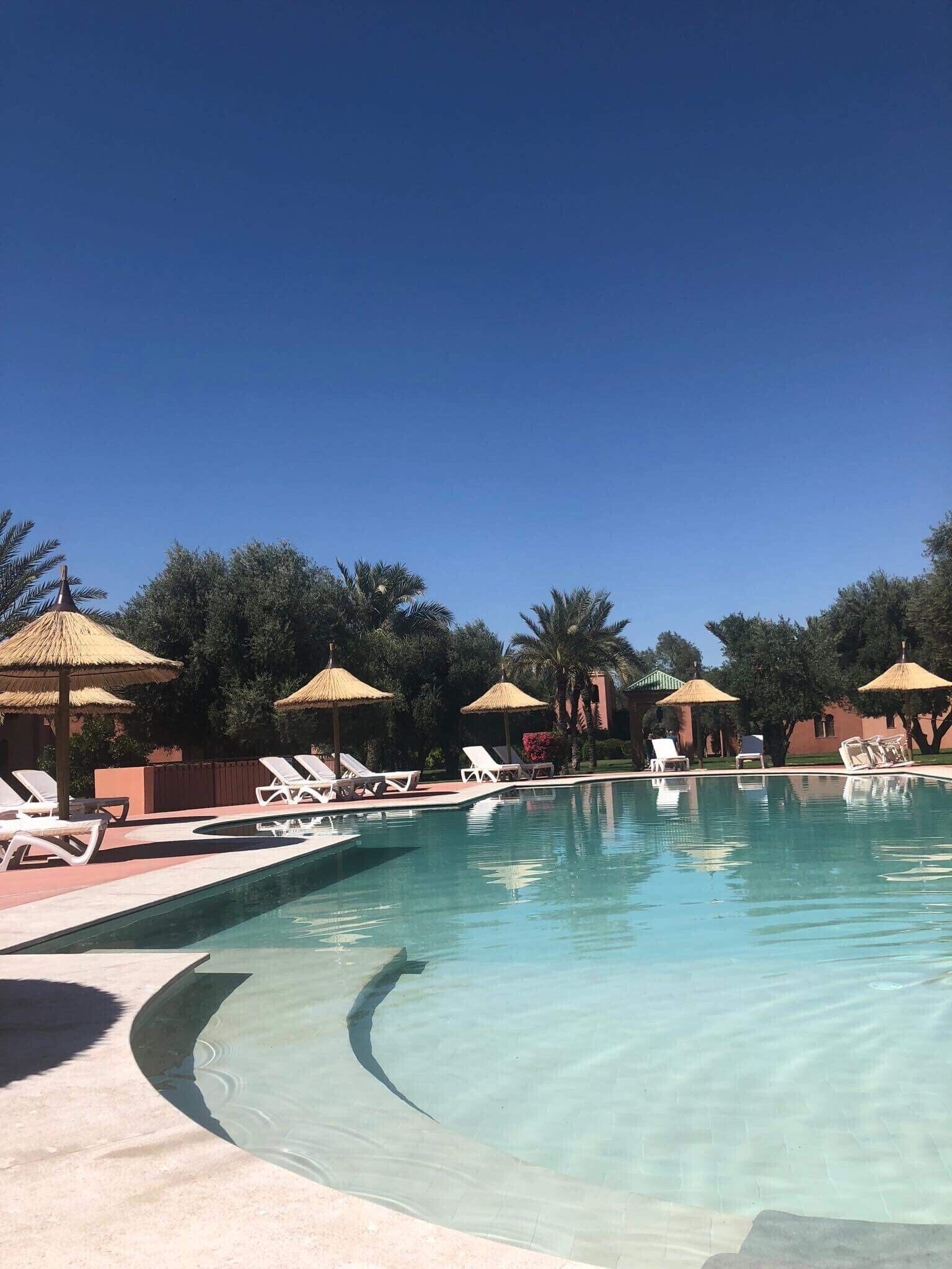 Una piscina cristalina con sillas de descanso y sombrillas de paja, rodeada de árboles y un cielo despejado.