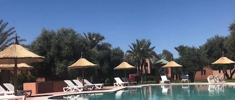 Una piscina cristalina con sillas de descanso y sombrillas de paja, rodeada de árboles y un cielo despejado.