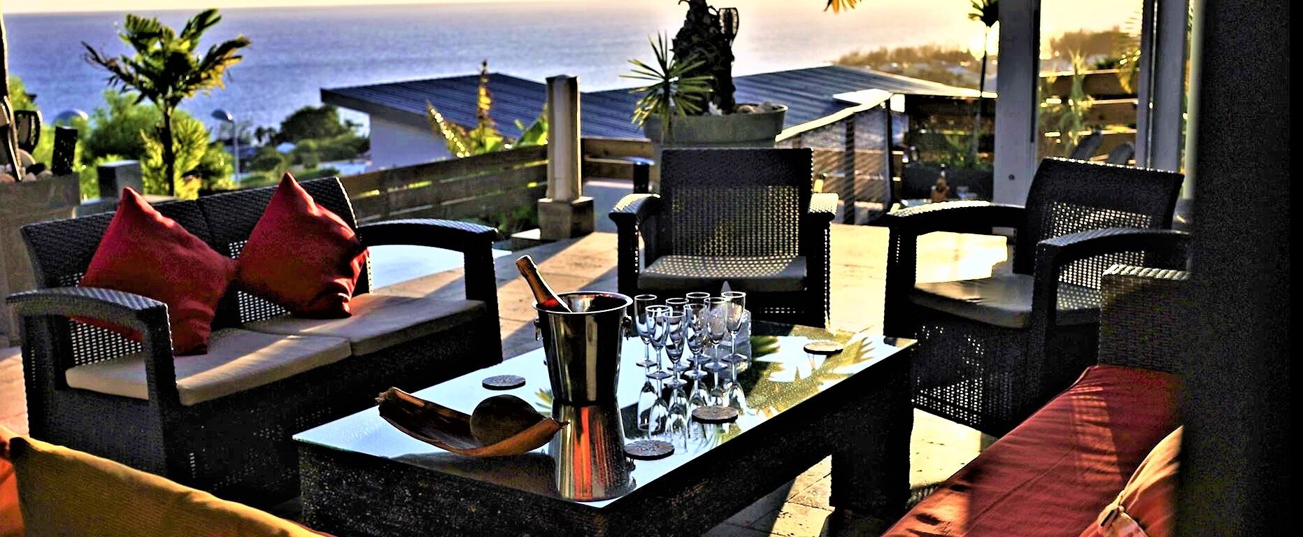 Un espace extérieur aménagé avec des chaises et des coussins en osier, une carafe en métal et des verres à vin posés sur une table.