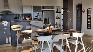 A modern kitchen with a glass dining table and white chairs, a built-in oven, and a refrigerator.