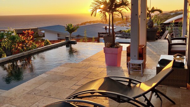 A patio with a pool, lounge chairs, and a view of the ocean at sunset.
