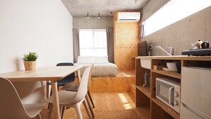 A compact, modern room with a wooden floor, a small dining table with chairs, a bed, a kitchenette with a sink and stove, and a window with curtains. - Smi:re Stay Oshiage (Tokyo)