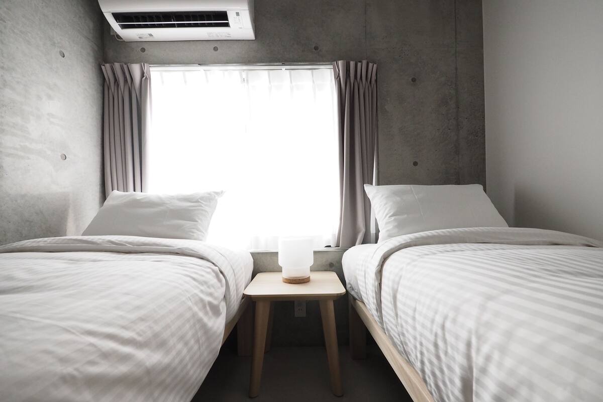 A modern hotel room with two single beds, a small wooden nightstand, a window with curtains, and an air conditioning unit.