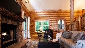 L’intérieur d’un chalet en bois présente une cheminée en pierre, un canapé en cuir et une grande fenêtre donnant sur le paysage extérieur.