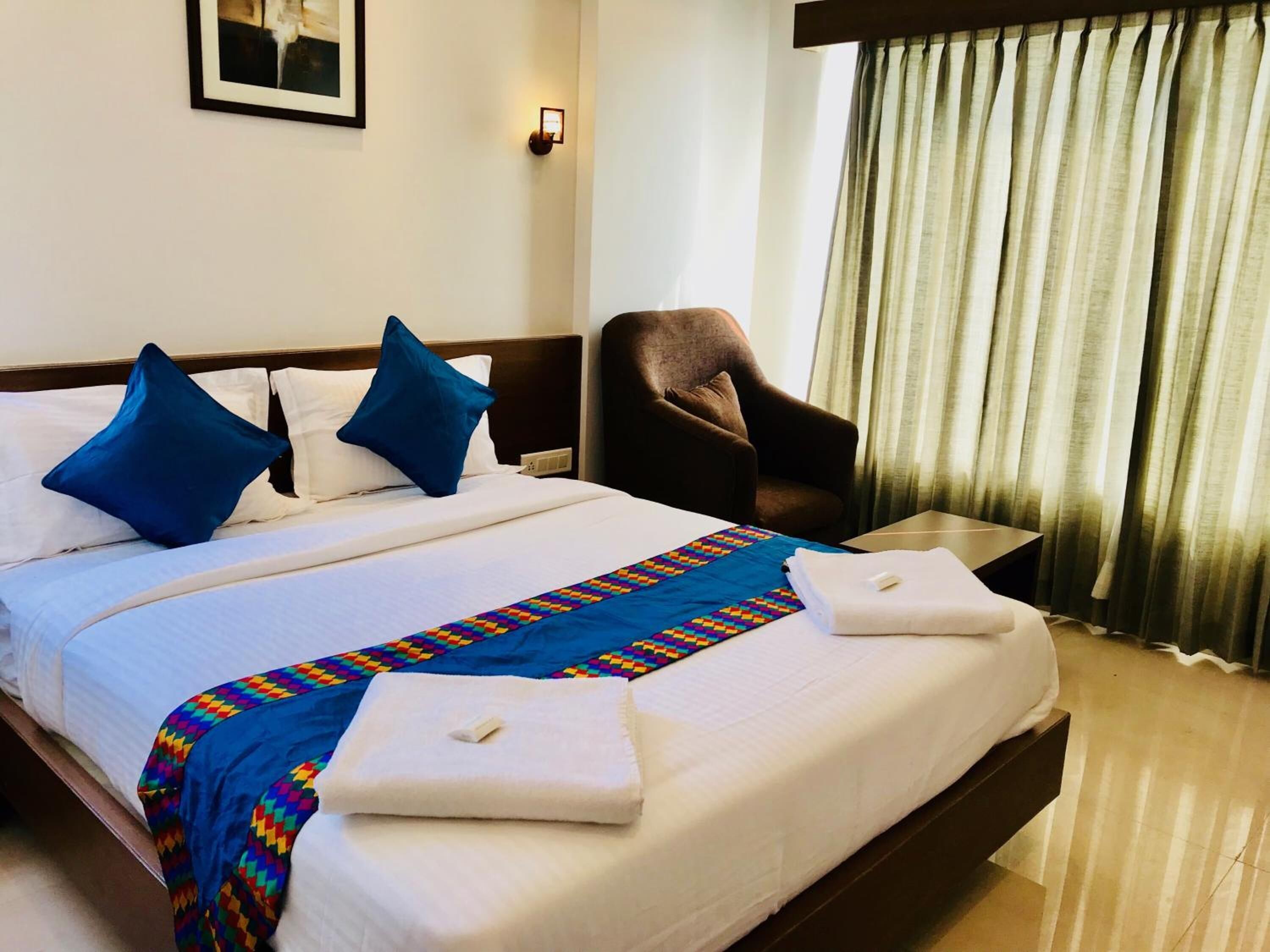 Quarto de hotel com cama bem arrumada, uma cadeira, uma mesinha e uma janela com cortinas.