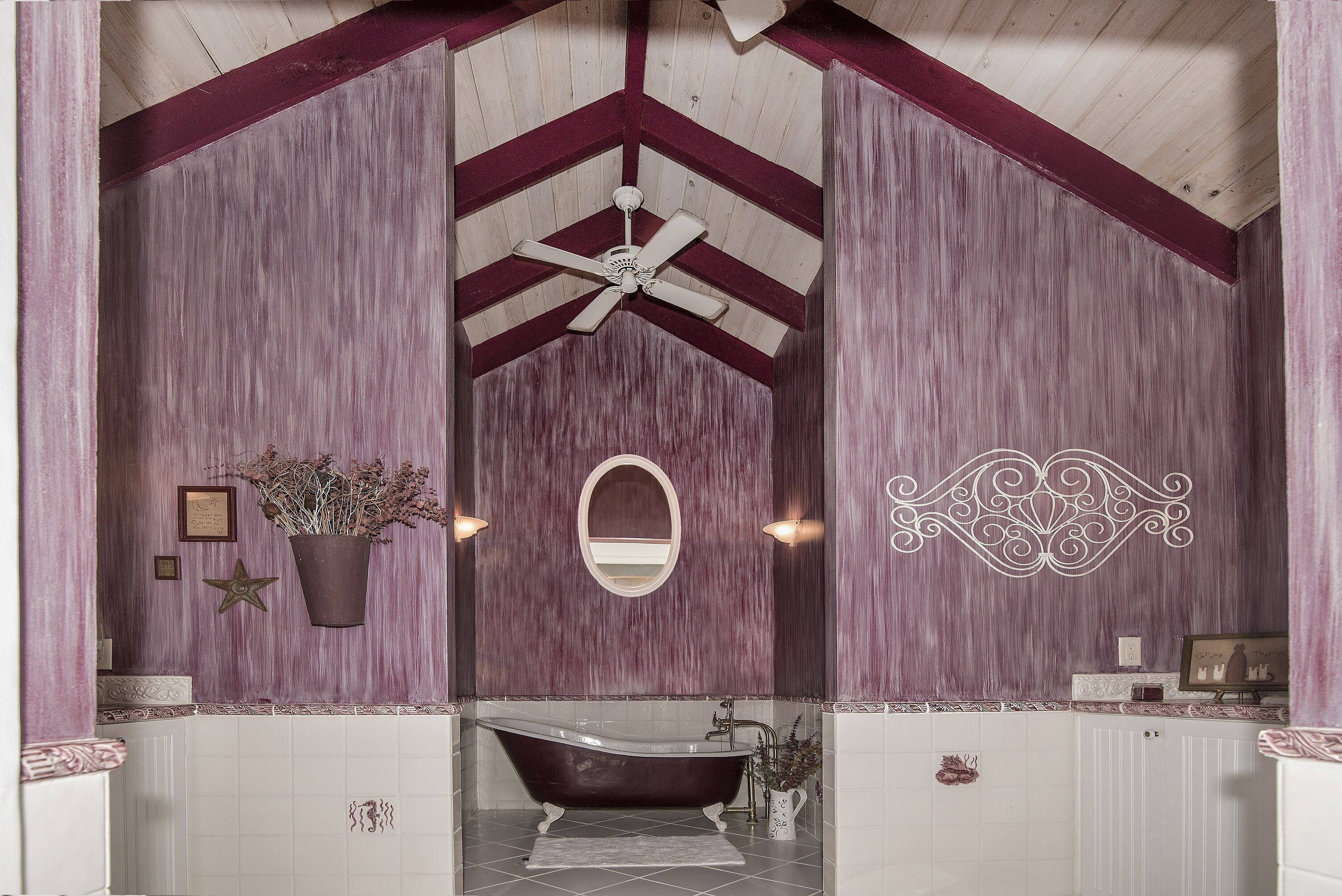 Casa de banho com banheira de pé, ventoinha no teto, espelho e detalhes decorativos na parede.