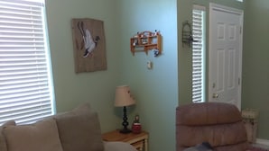 Ein Wohnzimmer mit einem beigefarbenen Sofa, einem Holztisch und einem braunen Sessel. Es gibt ein kleines Holzregal mit Dekoration und eine Lampe auf einem Beistelltisch.