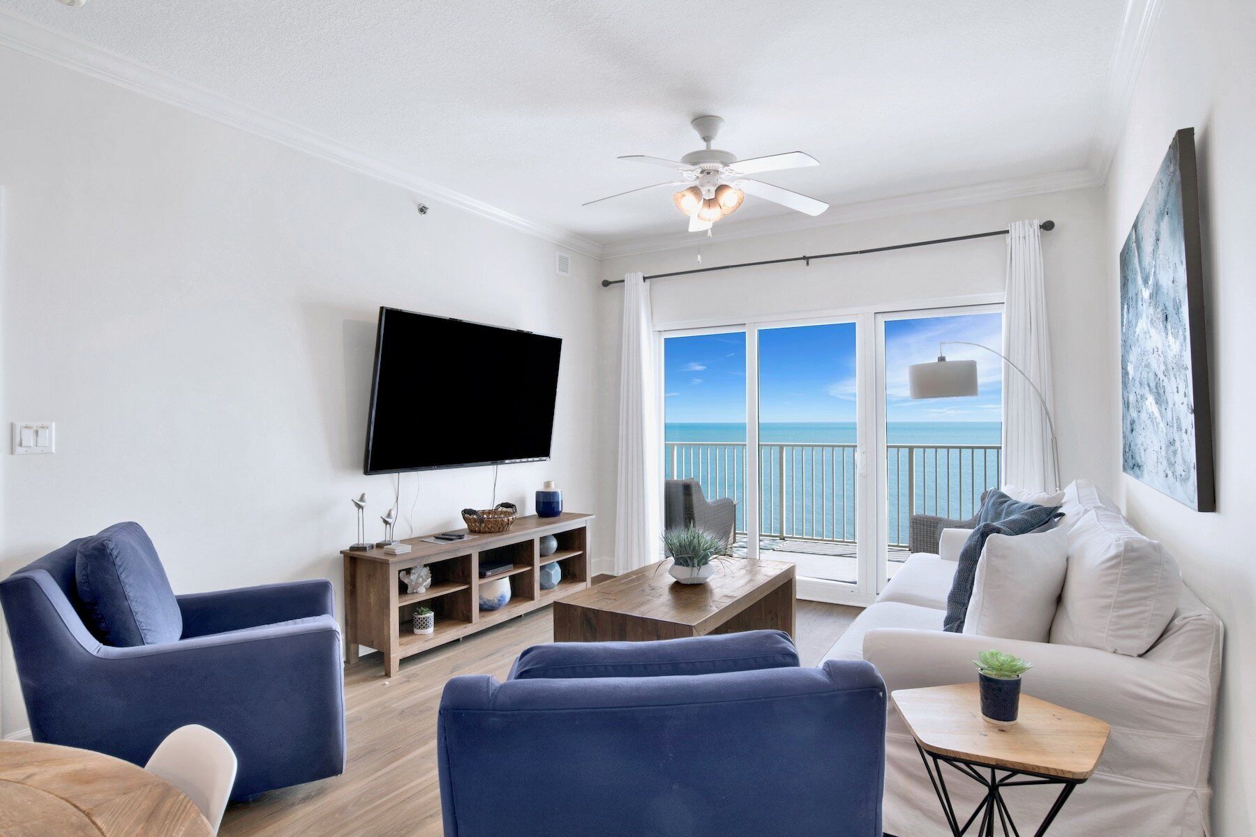A living room with a flat-screen TV, blue sofas, a wooden coffee table, and a balcony with a view of the ocean.