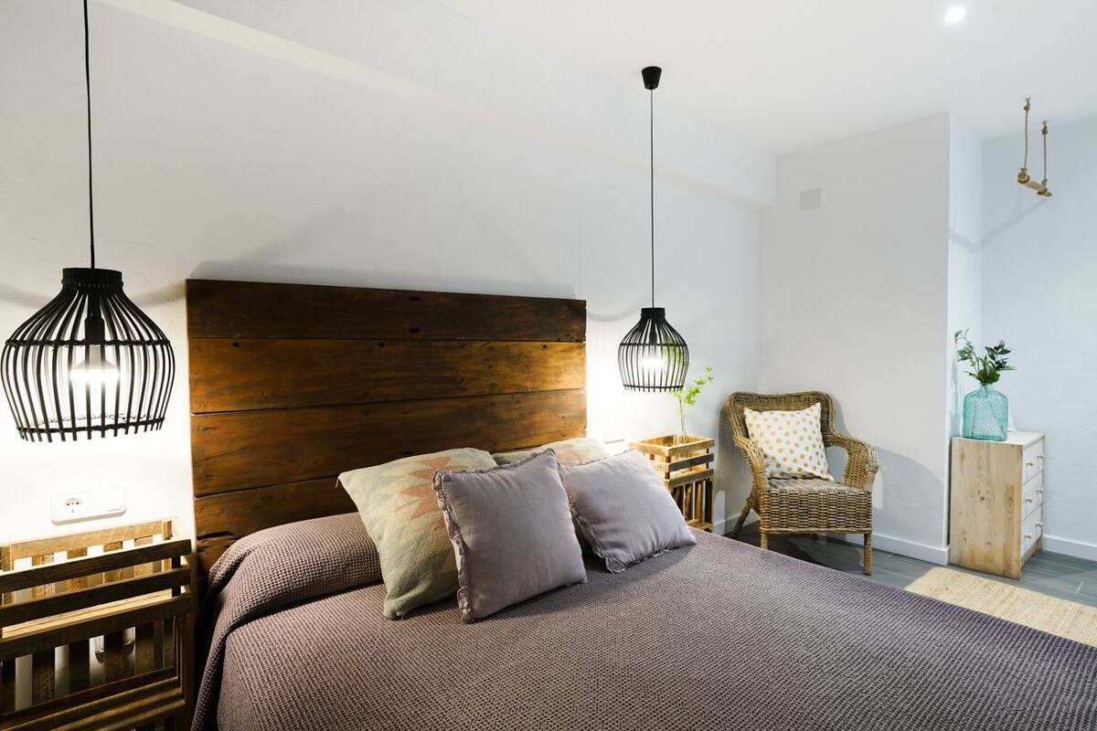 Un dormitorio con cabecera de madera, dos lámparas colgantes, un sillón de mimbre, una mesita de noche de madera y una planta.