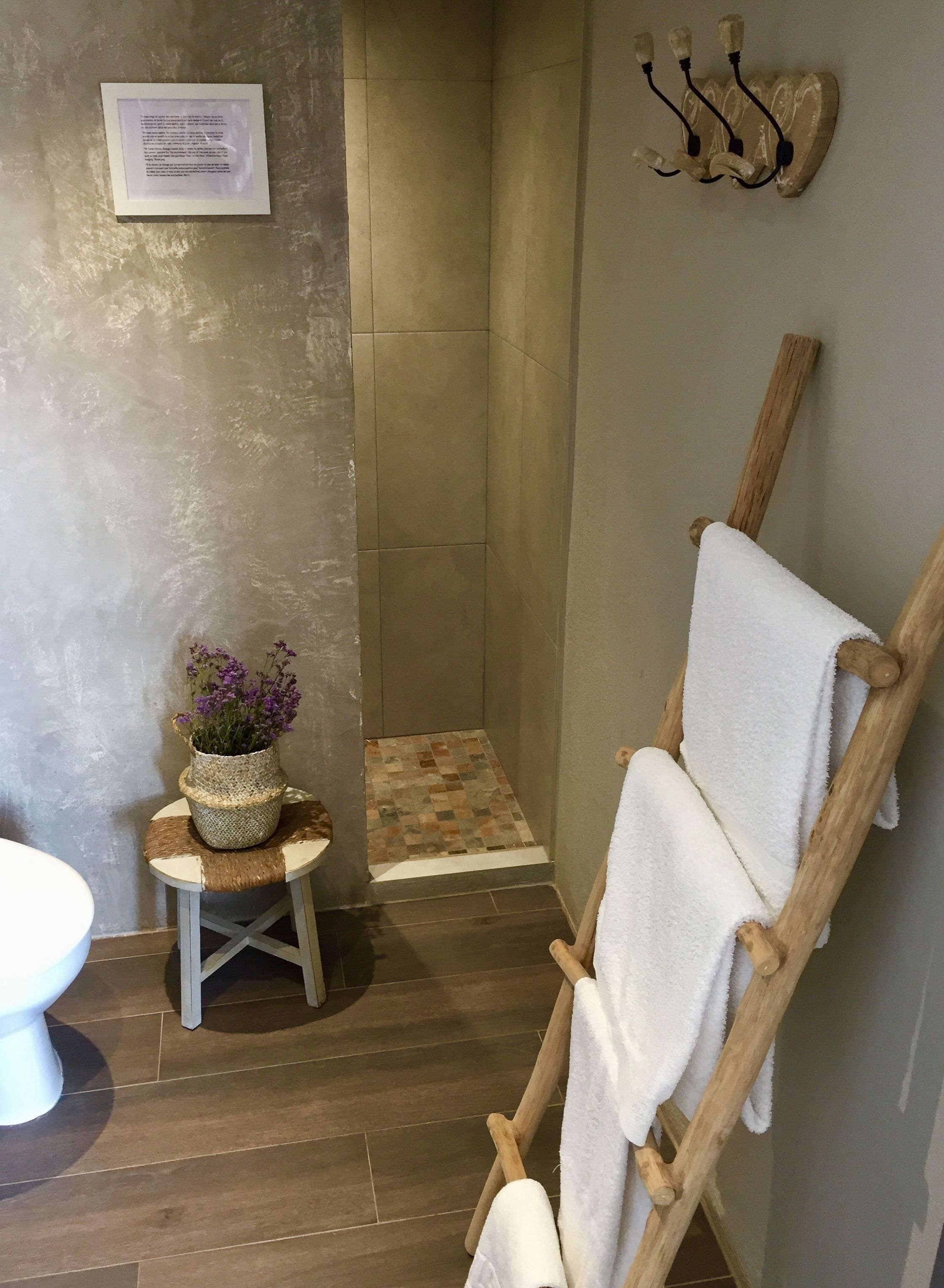 A bathroom with a wooden towel rack, a small stool with a woven basket of flowers, a shower area, and a sign on the wall.