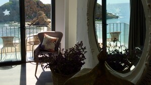 Habitación con un ventanal grande que da a un litoral rocoso y al mar. Se ve una silla de mimbre con cojín y una planta en maceta en primer plano.