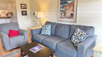 A living room with a grey sofa, a red pillow, a coffee table with a book, a lamp, and a framed painting on the wall.