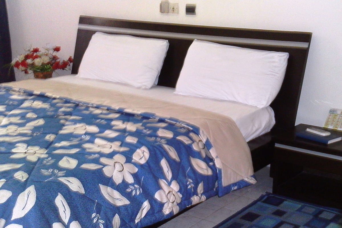 A bed with white pillows and a blue floral bedspread, a nightstand with a book and remote, and a small vase with flowers on the left.