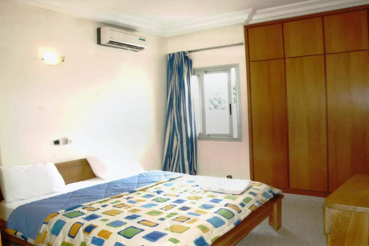 A bedroom with a bed, a wooden wardrobe, a window with curtains, and an air conditioning unit.