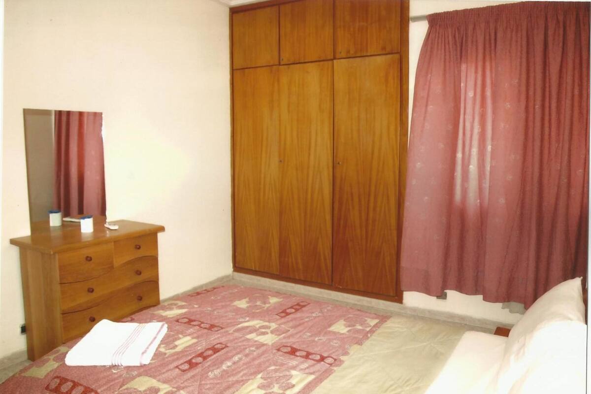 A bedroom with a wooden wardrobe, a bed with a patterned bedspread, and a mirror on a dresser.