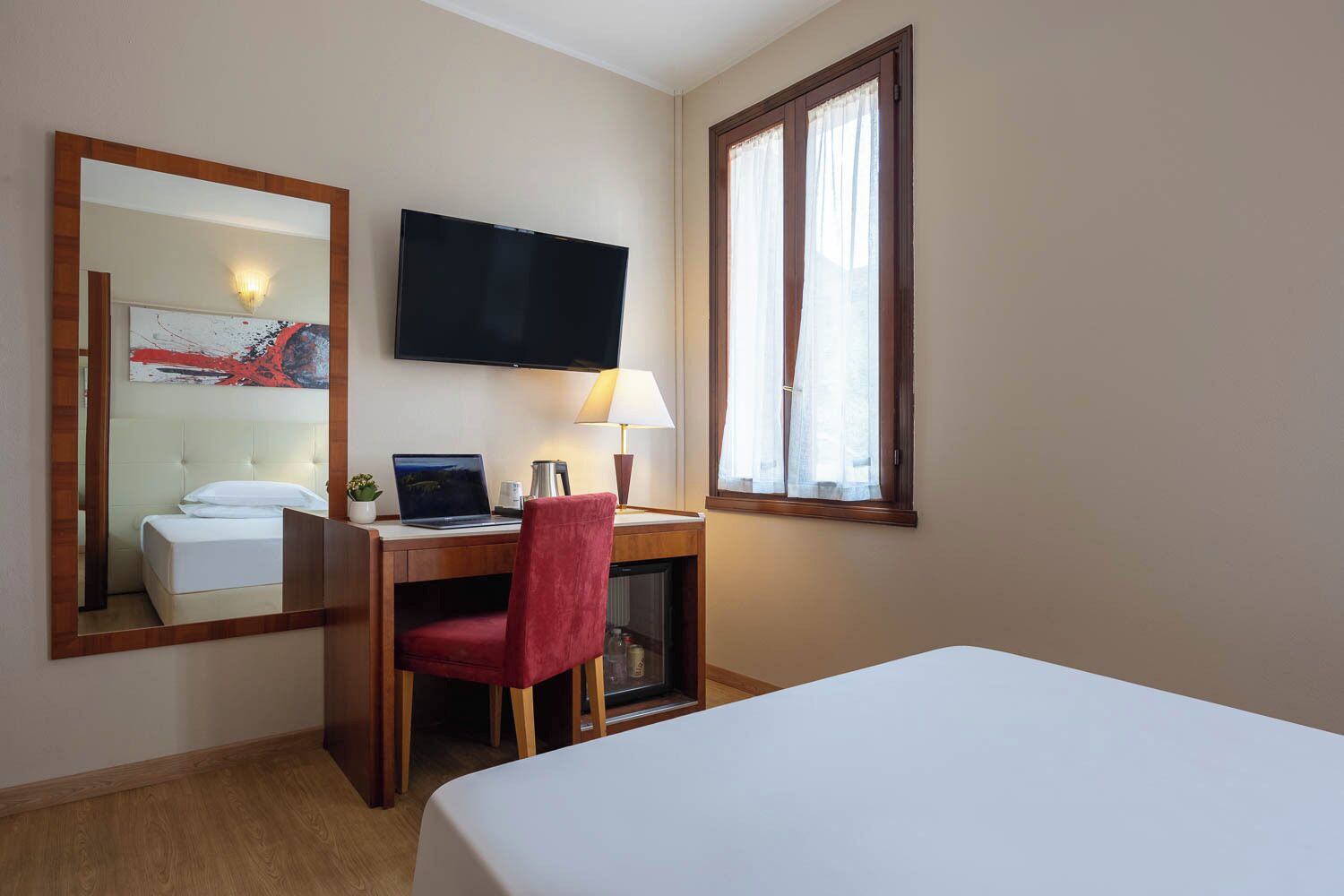 Habitación de hotel con cama, escritorio con computadora portátil, silla, televisor y ventana con cortinas.