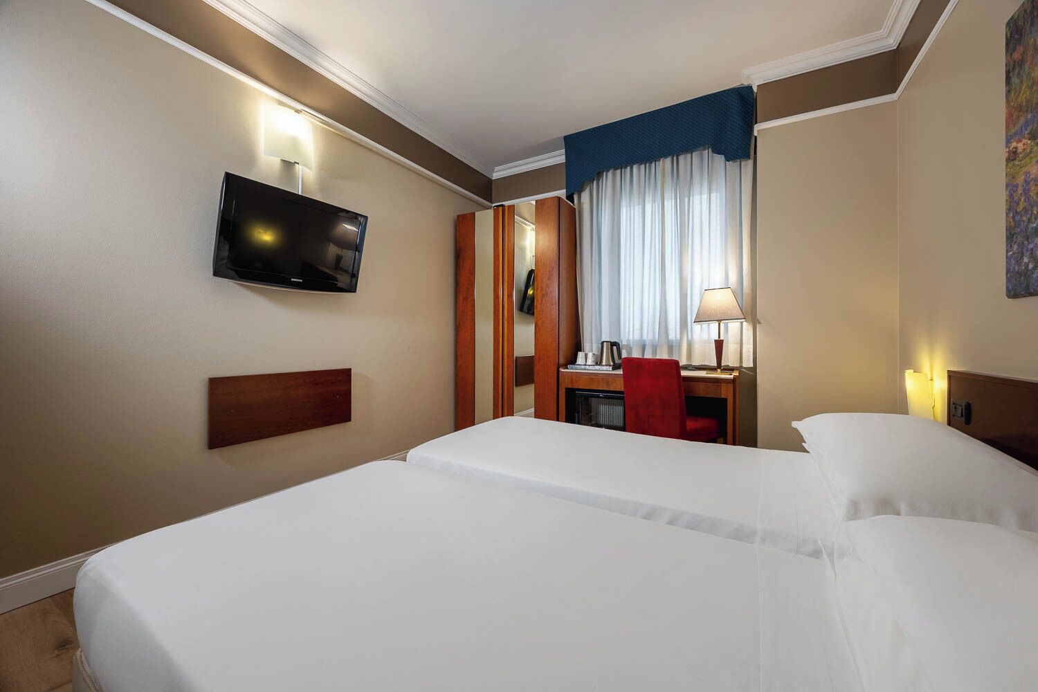 Une chambre d’hôtel avec deux lits, un téléviseur fixé au mur, un bureau avec une chaise et une fenêtre avec des rideaux.