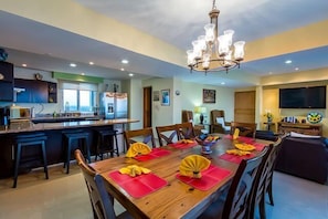 A modern dining area with a wooden table set for six, surrounded by chairs. A chandelier hangs above the table, and the room features a kitchen area with dark cabinetry and a bar. - Large Condo, Heated Pool, Sauna, Massage Room (Cozumel)