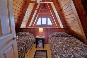 A cozy attic bedroom with wooden walls, a sloped ceiling, two beds with patterned bedding, a small table with a lamp, and a window providing natural light.